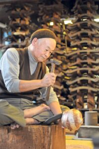 An elderly Japanese craftsman hammering a metal kettle in his workshop.
